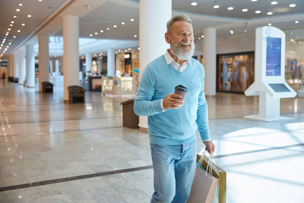 Przystojny, elegancki staruszek z torbami na zakupy i filiżanką kawy w centrum handlowym. Koncepcja dnia zakupów - Zdjęcie, obraz