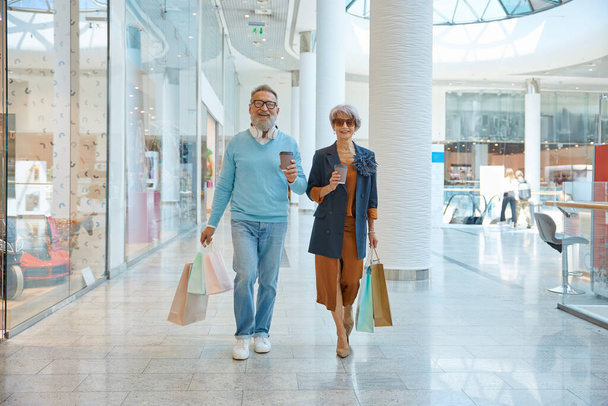 Fashionable elderly couple enjoying shopping tour on weekends. Happy aged man and woman on retirement choosing new trendy outfit in city mall - Photo, Image