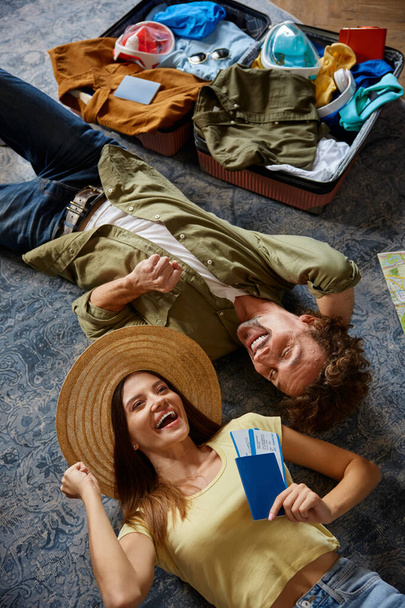 Happy man and woman couple traveler lying on floor nearby opened suitcase with belongings cheering to buy tour with sale discount. Summer holiday vacation trip planning concept - Photo, Image