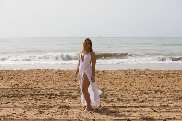 Jovem mulher loira bonita em vestido branco está andando na areia na costa da praia em um dia ensolarado. A mulher faz diferentes expressões corporais. No fundo o mar azul. - Foto, Imagem