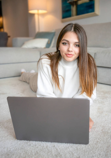 Ładna brunetka używająca laptopa leżącego na podłodze w salonie. Pracuje w domu w zamknięciu kwarantanny. Samoizolacja na dystans społeczny - Zdjęcie, obraz