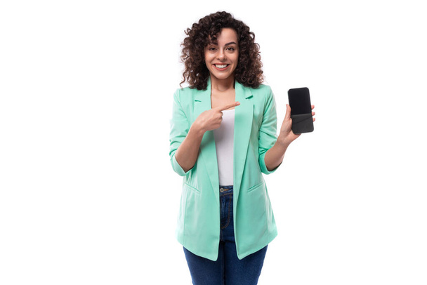 νεαρό λεπτό καυκάσιο γυναικείο μοντέλο με σγουρά μαύρα μαλλιά δείχνει smartphone κάθετα με οθόνη mockup. - Φωτογραφία, εικόνα