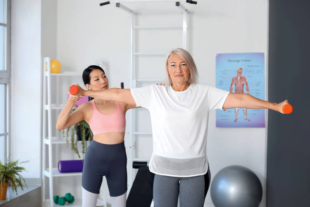 Mujer madura entrenando con pesas y terapeuta en centro de rehabilitación - Foto, Imagen