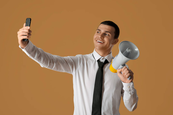 Молодой бизнесмен с мегафоном делает селфи на коричневом фоне - Фото, изображение