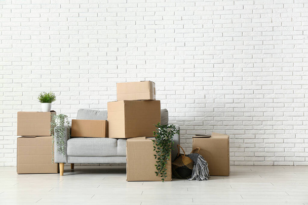 Диван с картонными коробками и растениями возле стены из белого кирпича в номере в день переезда - Фото, изображение