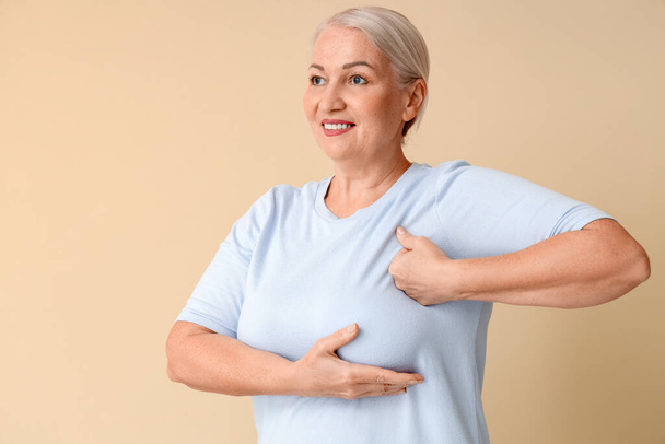 Reife Frau überprüft ihre Brust auf beigem Hintergrund. Krebsaufklärungskonzept - Foto, Bild