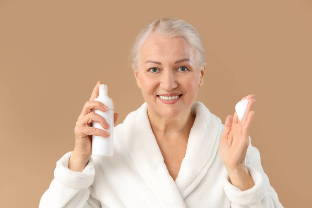 Beautiful mature woman with bottle of cleansing foam on brown background, closeup - Photo, Image