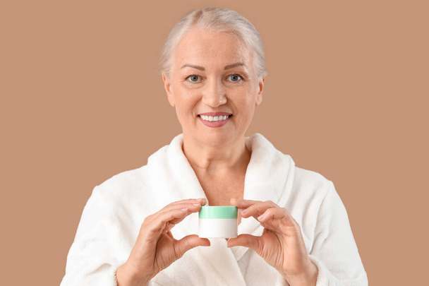 Beautiful mature woman with jar of cream on brown background, closeup - Photo, Image