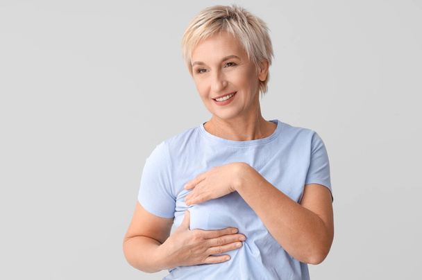 Lächelnde reife Frau überprüft ihre Brust auf hellem Hintergrund. Krebsaufklärungskonzept - Foto, Bild