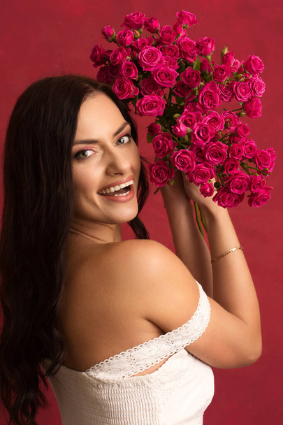 Beautiful young brunette woman with a bouquet of pink small roses on a red background - Photo, Image