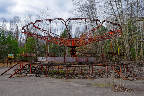 A rusted, abandoned carousel surrounded by overgrown trees and a dilapidated building. - Photo, Image