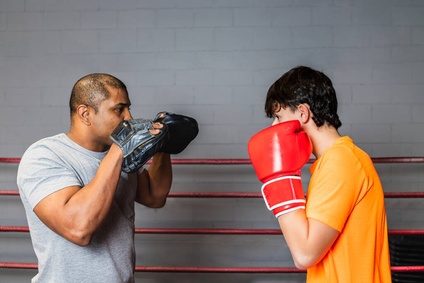 Horizontale foto blanke tiener jongen met oranje t-shirt en rode bokshandschoenen, in de ring met zijn mid volwassen zwarte trainer, leren boksen. Concept sport, recreatie. - Foto, afbeelding