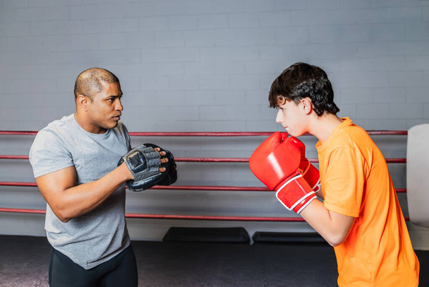 Горизонтальное фото белого мальчика-подростка в красных боксерских перчатках и оранжевой футболке, со своим средним взрослым тренером по черному боксу, на ринге. Концепция спорта, отдыха. - Фото, изображение