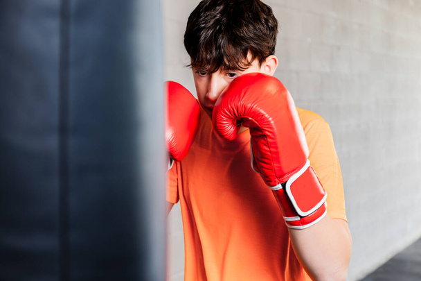 Foto orizzontale ragazzo adolescente caucasico con t-shirt arancione e guanti da boxe rossi, in una palestra di pugilato. Ricevuto. Concetto sport, ricreazione. - Foto, immagini