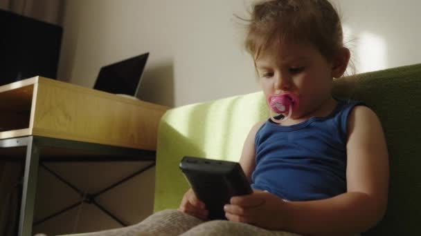 Кавказская малышка подлизывается к пустышке, используя смартфон, играя в игры на диване дома. Концепция образа жизни с использованием детских технологий. - Кадры, видео