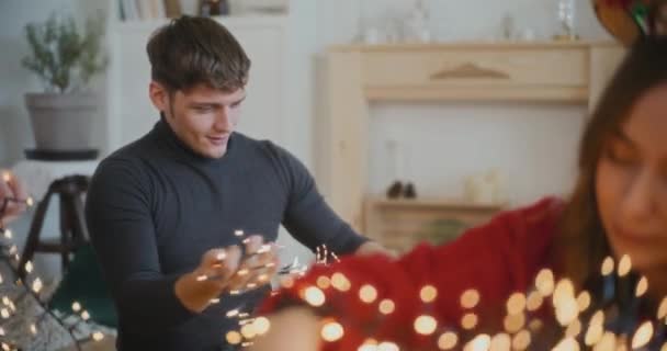Молодой человек сортирует запутанные светильники с подружками дома на Рождество - Кадры, видео