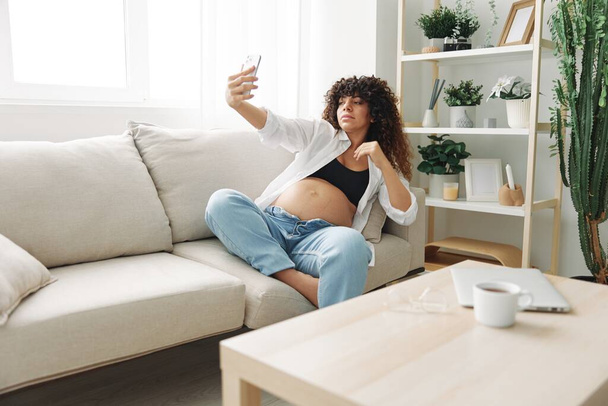Hamile kadın blogcu evdeki kanepede oturur ve telefonda kendi fotoğraflarını çeker, selfie ve video görüşmesi, doktorla online istişare, hamilelik yönetimi. Yüksek kalite fotoğraf - Fotoğraf, Görsel