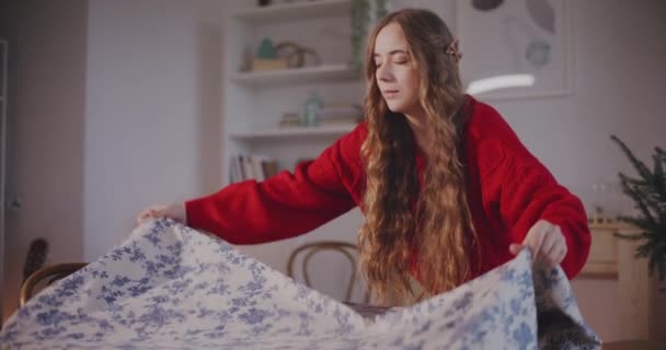 Giovane donna bionda mettere tovaglia sul tavolo mentre in piedi in soggiorno - Filmati, video