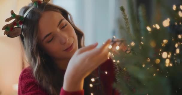 Femme souriante ajustant les lumières LED éclairées sur l'arbre à la maison pendant les vacances de Noël - Séquence, vidéo