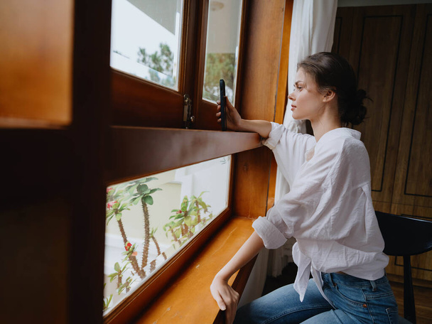 Beautiful woman with phone in hand sitting at the window with wooden frame of the house, home comfortable lifestyle with online work, cozy atmosphere and aesthetics, spring time. High quality photo - Photo, Image