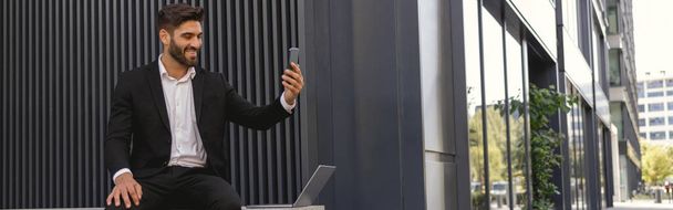 Lächelnder Geschäftsmann macht Selfie mit Handy auf Bank in der Nähe von Bürogebäude - Foto, Bild
