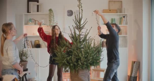 Hombres y mujeres jóvenes ajustando luces en el árbol de Navidad en casa durante las vacaciones - Metraje, vídeo