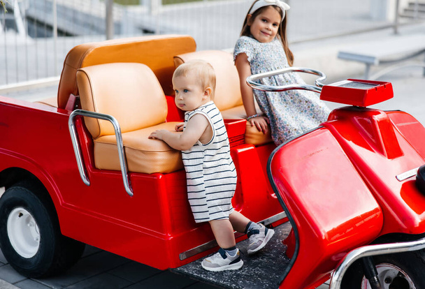 Un niño y una niña juegan en un parque público y montan en un coche rojo de juguete. Los niños pequeños pasan su tiempo libre al aire libre en el parque. Hermano pequeño y su hermana mayor en un coche rojo para niños - Foto, imagen