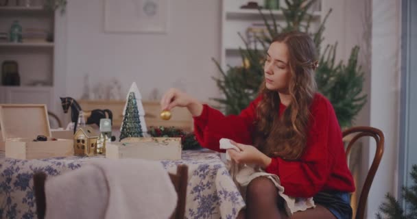 Young woman cleaning bauble with cloth while sitting on chair in living room at home - Footage, Video