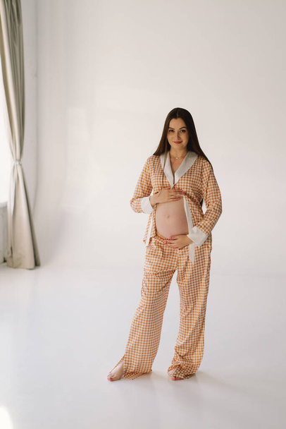 Милая молодая беременная девушка сидит на белом студио в теплом свете с руками на беременном желудке на фоне стен с тенью. Беременная концепция, свободное пространство, наслаждение беременностью. Беременное тело. - Фото, изображение