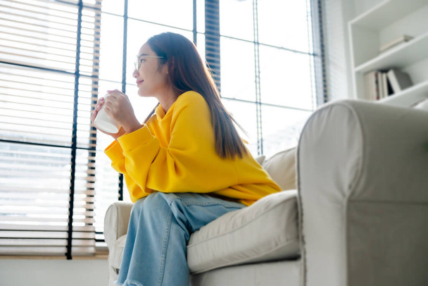 Młoda Azjatka w okularach, siedząca na kanapie i trzymająca kubek w salonie, pijąca rano mleko lub herbatę. Zdrowy i odświeżony nastrój - Zdjęcie, obraz