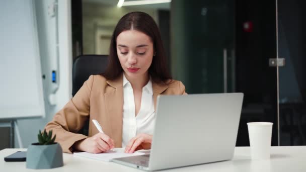 Busy caucasian entrepreneur typing on portable laptop and noting information on piece of paper in office corporate. Confident woman writing information about business project in notebook at workplace. - Footage, Video
