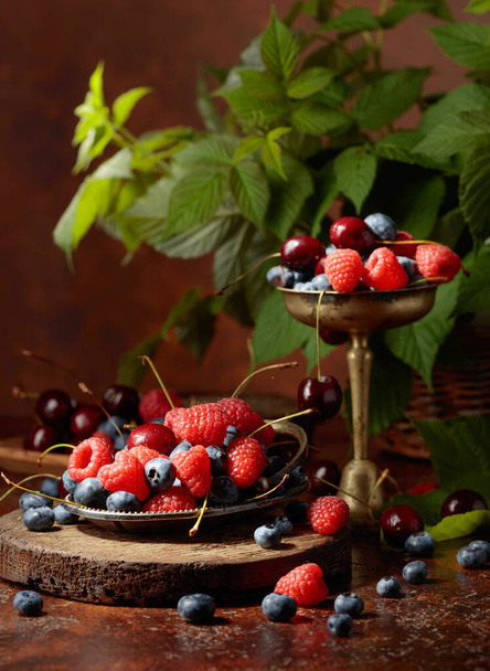 Ягоды с листьями на старом коричневом столе. Разноцветная смесь черники, малины и вишни. - Фото, изображение