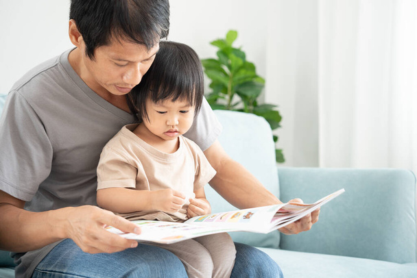 Mutlu Asyalı baba rahatla ve evde bebek zamanlarıyla kitap oku. Ebeveyn kızıyla kanepede oturup masal okuyor. öğrenme, çocuk bakımı, gülme, eğitim, hikaye anlatma, uygulama. - Fotoğraf, Görsel