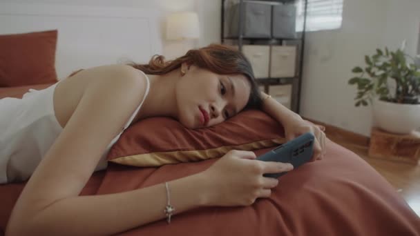 Handheld shot młodej zdenerwowanej kobiety za pomocą smartfona i czytania wiadomości podczas odpoczynku w łóżku sam w domu - Materiał filmowy, wideo