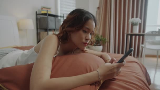 Handheld laukaus väsynyt aasialainen tyttö kutoma viestejä matkapuhelimeen, kun makaa helmi kotona pitkän päivän jälkeen töissä - Materiaali, video