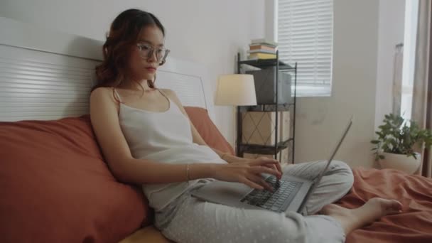 Handheld laukaus mietteliäs naispuolinen opiskelija tai freelancer työskentelee langattoman tietokoneen istuu sängyssä kodikas huone - Materiaali, video