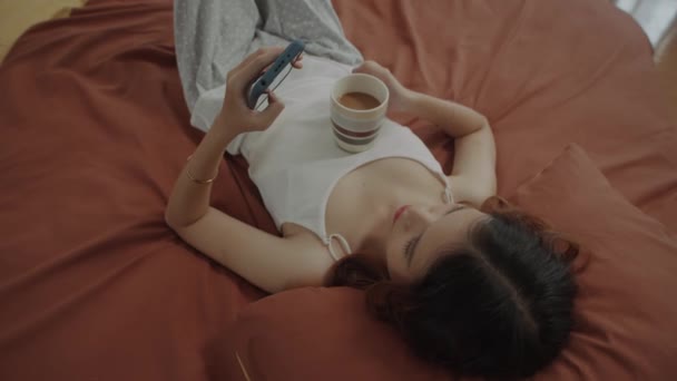 Direkt über der Ansicht einer Frau, die Nachrichten auf dem Handy checkt, während sie es sich zu Hause im Bett gemütlich macht - Filmmaterial, Video