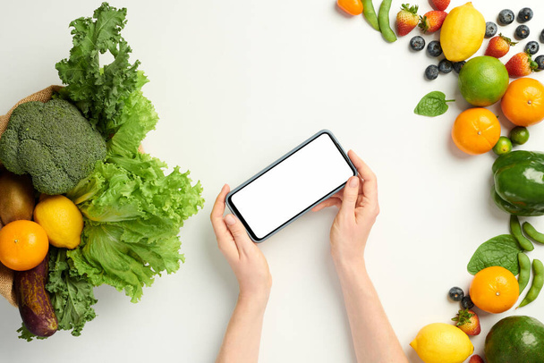 Mani della persona che tiene smartphone con schermo bianco sopra il tavolo con generi alimentari biologici freschi dal supermercato - Foto, immagini