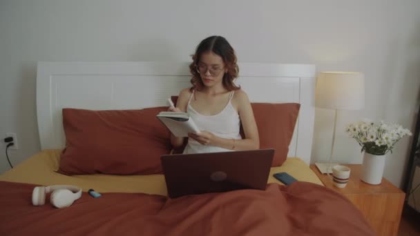 Середній довгий знімок студентки робить нотатки, сидячи перед ноутбуком в ліжку під час домашнього навчання - Кадри, відео