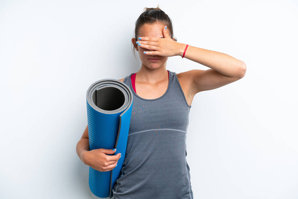 Young sport woman going to yoga classes while holding a mat isolated on white background covering eyes by hands. Do not want to see something - Photo, Image