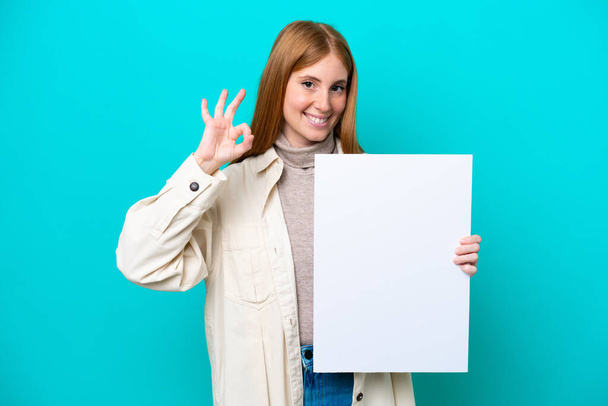 Junge rothaarige Frau isoliert auf blauem Hintergrund hält ein leeres Plakat und tut OK Zeichen - Foto, Bild