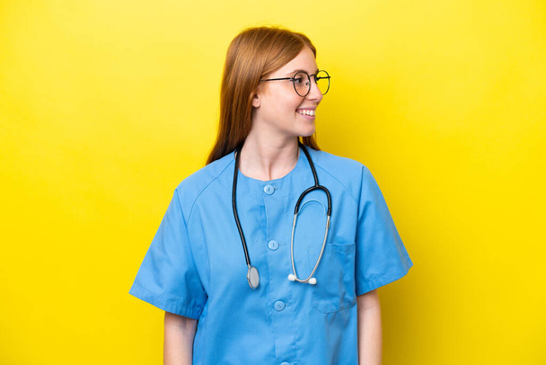 Mladý ryšavý zdravotní sestra žena izolovaný na žlutém pozadí hledá straně - Fotografie, Obrázek