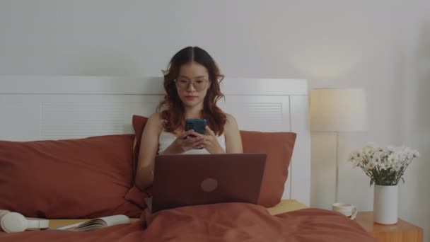 Media colpo lungo di donna asiatica sms messaggio al collega mentre si lavora da casa e utilizzando il computer portatile seduto sul letto - Filmati, video