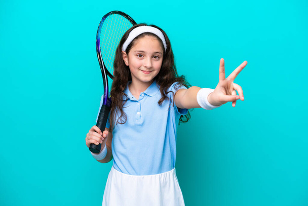 Pieni valkoihoinen tyttö pelaa tennistä eristetty sinisellä taustalla hymyillen ja osoittaa voiton merkki - Valokuva, kuva