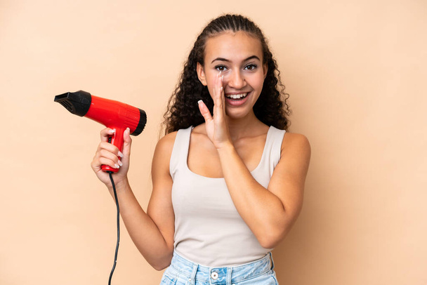 Young woman holding a hairdryer isolated on beige background whispering something - Photo, Image