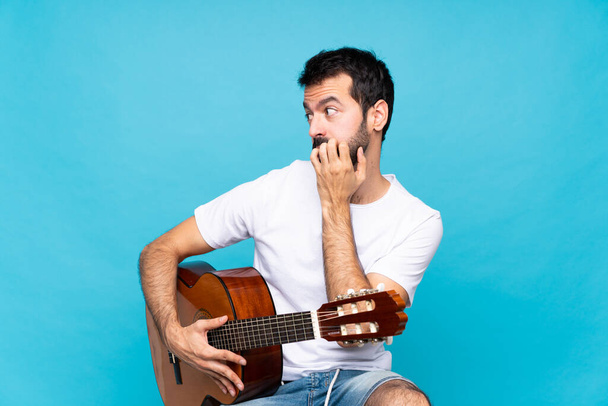 孤立した青い背景の上にギターを持つ若い男は緊張し,口に手を置くことを恐れました - 写真・画像