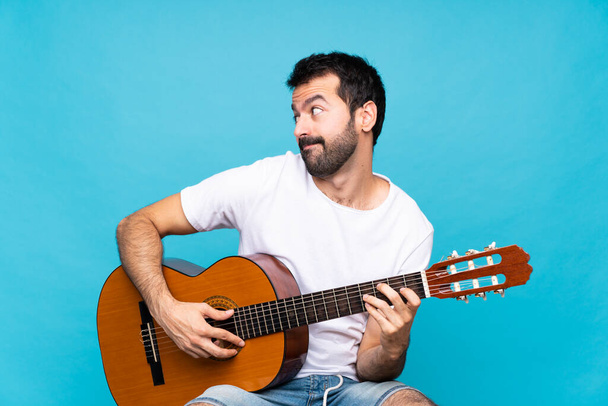 Fiatal férfi gitár felett elszigetelt kék háttér így kétséges gesztus látszó oldalon - Fotó, kép