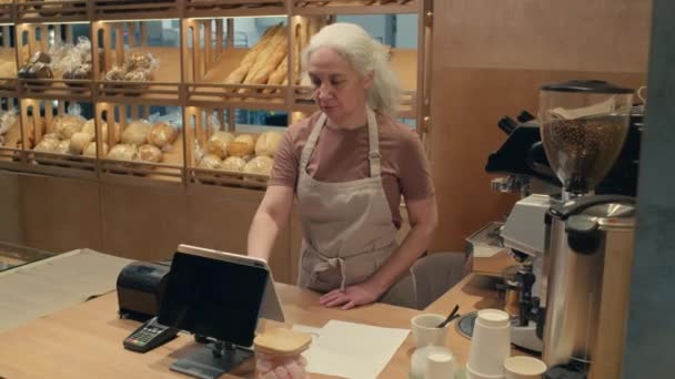 Senior vrouwelijke werknemer in schort met behulp van kassa monitor aan de balie tijdens de werkdag in de bakkerij - Video