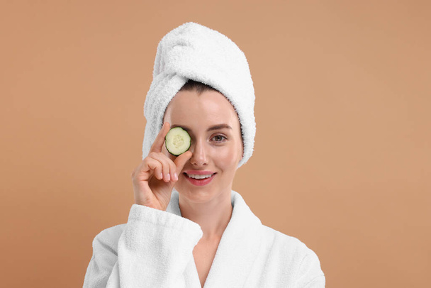 Beautiful woman in bathrobe covering eye with piece of cucumber on beige background - Фото, зображення