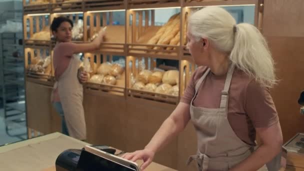 Tilt up shot van senior kassier werken aan de balie en vragen jonge vrouwelijke collega voor gehouden tijdens de dag in de bakkerij - Video